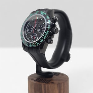 May include: A black chronograph wristwatch featuring a black dial with three sub-dials, a red second hand, and a tachymeter bezel with green numerals. The watch has a black case and a black silicone strap, displayed on a wooden stand.