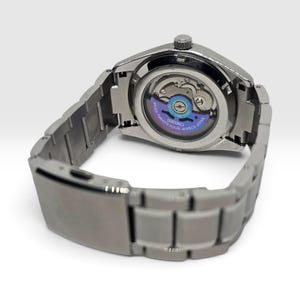 May include: A silver-toned stainless steel wristwatch with a metallic link bracelet and a deployment clasp. The watch features an exhibition case back revealing the internal mechanical movement, with a purple rotor marked Seiko 4R36A Twenty-Four Jewels.