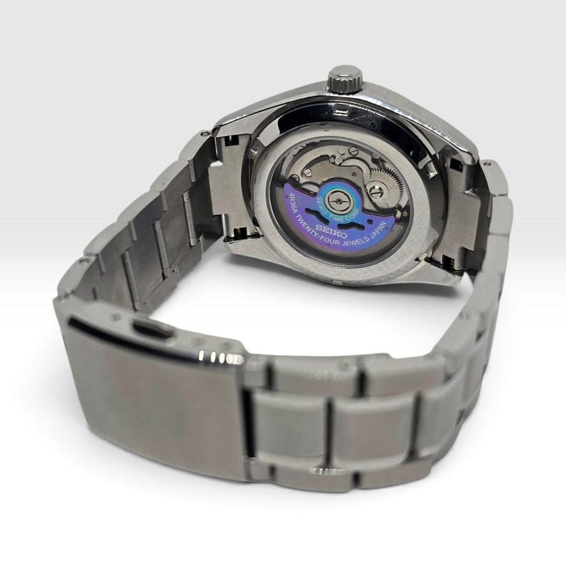 May include: A silver-toned stainless steel wristwatch with a metallic link bracelet and a deployment clasp. The watch features an exhibition case back revealing the internal mechanical movement, with a purple rotor marked Seiko 4R36A Twenty-Four Jewels.
