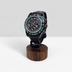 May include: A black chronograph wristwatch with a dark gray dial, three sub-dials, and a tachymeter bezel featuring green text. The watch has a black silicone strap and is mounted on a cylindrical wooden display stand with the text SQND engraved.