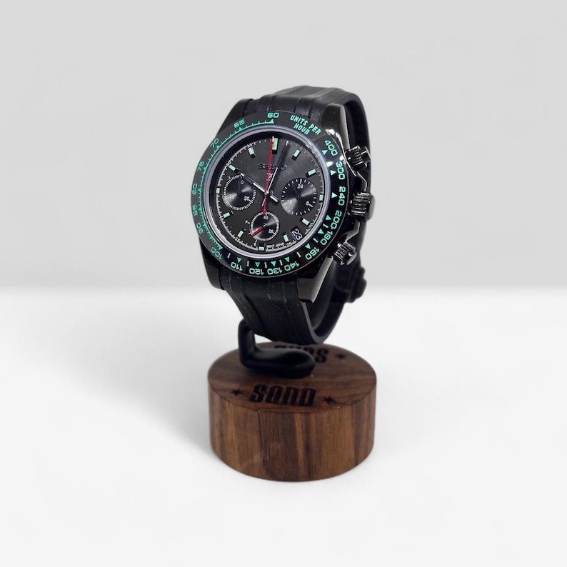 May include: A black chronograph wristwatch with a dark gray dial, three sub-dials, and a tachymeter bezel featuring green text. The watch has a black silicone strap and is mounted on a cylindrical wooden display stand with the text SQND engraved.