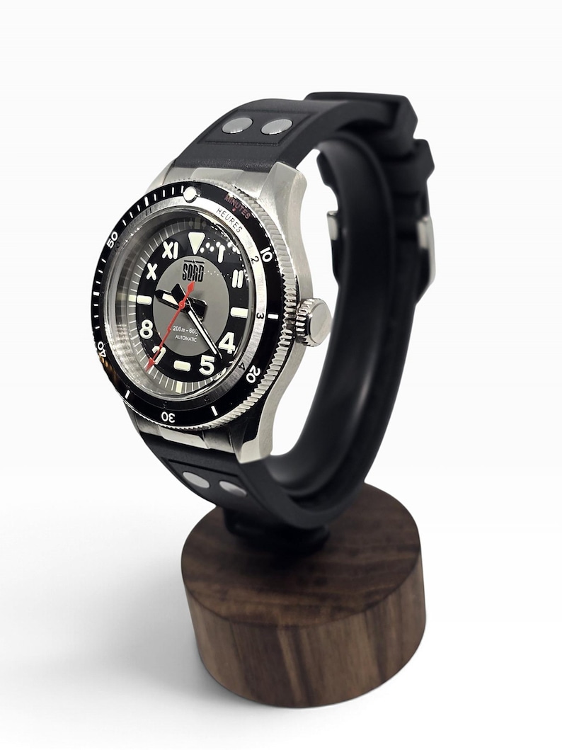 Puede incluir: Un reloj de pulsera autom&aacute;tico con correa de caucho negra que presenta dos remaches plateados. El reloj tiene una caja de acero inoxidable plateado, un bisel giratorio negro con marcas blancas y una esfera gris con el texto SORD, 200m-660ft y AUTOMATIC.