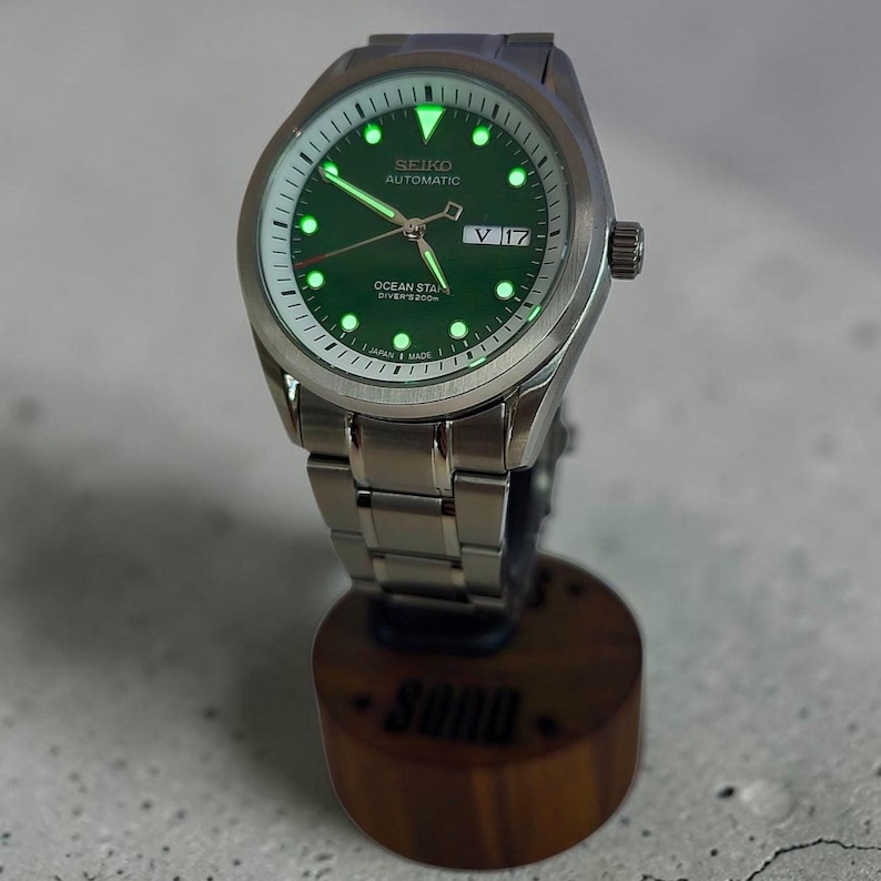May include: A stainless steel automatic wristwatch with a green dial, luminous hour markers, and hands. The watch face displays the text SEIKO AUTOMATIC, OCEAN STAR, DIVER'S 200M, and JAPAN MADE, with a day-date window showing V 17 on the right side.
