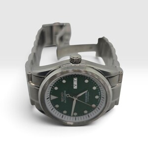 May include: A silver-tone stainless steel automatic wristwatch featuring a dark green dial with luminous hour markers, silver hands, and a day-date window at the three o'clock position. The watch face displays the text Seiko Automatic Ocean Star.