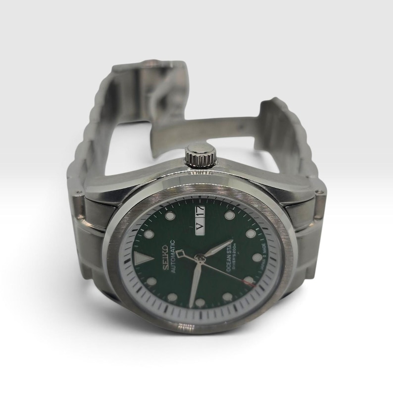 May include: A silver-tone stainless steel automatic wristwatch featuring a dark green dial with luminous hour markers, silver hands, and a day-date window at the three o'clock position. The watch face displays the text Seiko Automatic Ocean Star.