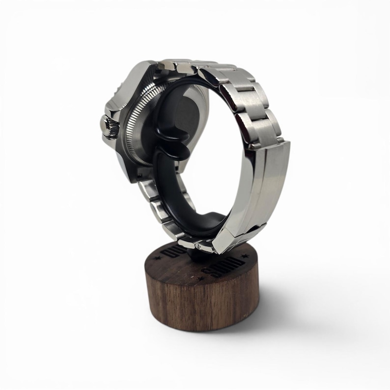 May include: A silver-toned metal wristwatch with a link bracelet mounted on a black curved holder, which is attached to a cylindrical dark brown wooden base featuring the engraved text "SDMD" and small star icons on its top surface.