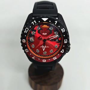 May include: A Seiko 5 automatic wristwatch featuring a black case and silicone strap. The dial is red with a black gradient, displaying white fist icons as hour markers, a yellow hat icon at the twelve o'clock position, and a date window.