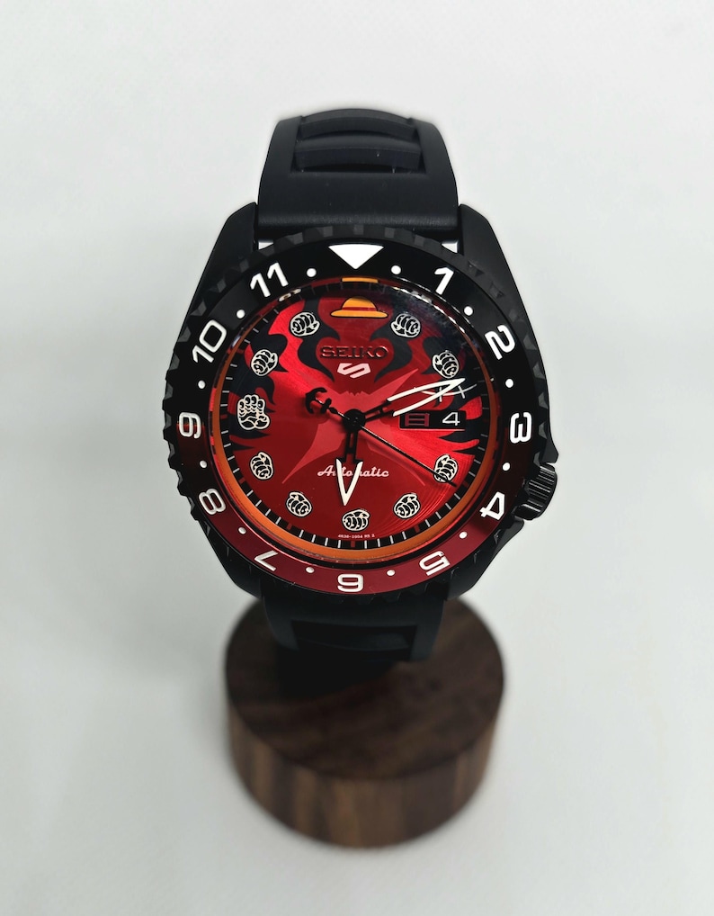 May include: A Seiko 5 automatic wristwatch featuring a black case and silicone strap. The dial is red with a black gradient, displaying white fist icons as hour markers, a yellow hat icon at the twelve o'clock position, and a date window.