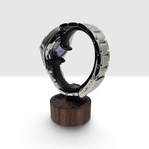 May include: A silver-toned wristwatch with a metallic link bracelet is displayed on a black curved holder mounted to a cylindrical dark wood base. The watch case back is visible, revealing internal mechanical components with a purple-tinted gear.