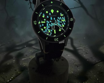 Puede incluir: Un reloj de pulsera Seiko con esfera negra que presenta un patrón luminoso de pequeñas figuras de fantasmas. El reloj tiene un bisel giratorio negro con números blancos, manecillas plateadas y una correa negra. Los marcadores brillan.