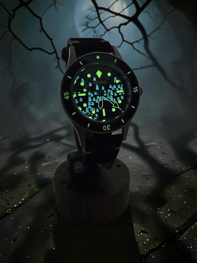 May include: A Seiko wristwatch with a black dial featuring a luminous pattern of small ghost figures. The watch has a black rotating bezel with white numerals, silver-tone hands, and a black strap. The dial markers and ghost shapes glow in the dark.