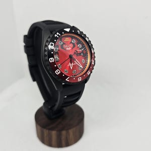 May include: A Seiko 5 automatic watch with a black silicone strap and a matte black case. The watch features a vibrant red dial with black graphic details, including a straw hat icon, white hour markers, and a date window at the three o'clock position.