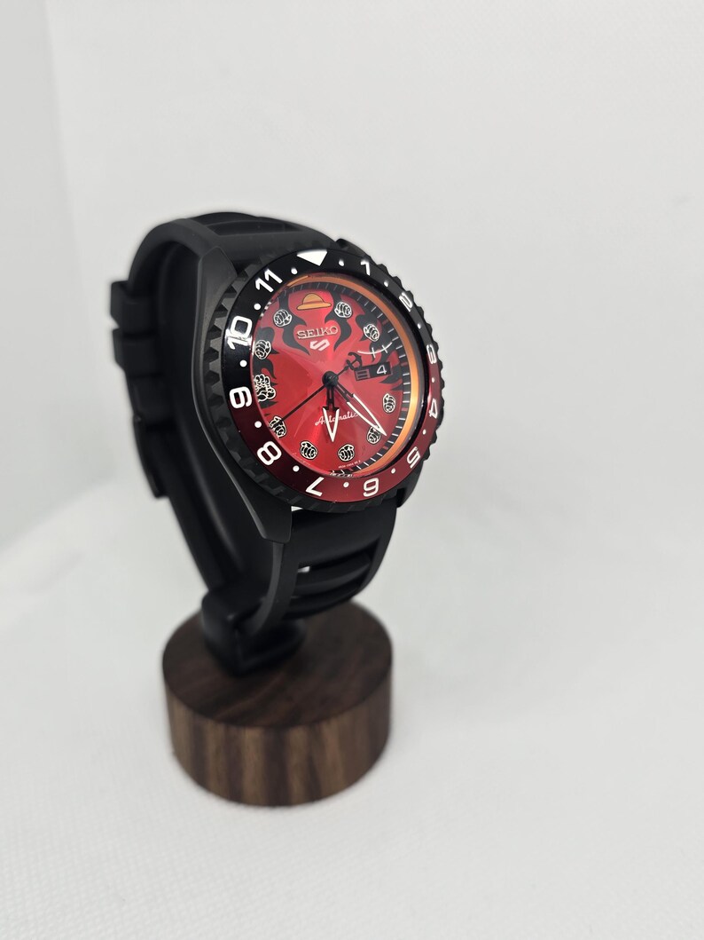 May include: A Seiko 5 automatic watch with a black silicone strap and a matte black case. The watch features a vibrant red dial with black graphic details, including a straw hat icon, white hour markers, and a date window at the three o'clock position.