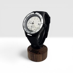 May include: An automatic wristwatch with a black case and a textured black rubber strap. The watch face features a silver-toned dial with a complex, map-like pattern, luminous hands, and a date window. The bezel is silver with black numerals.