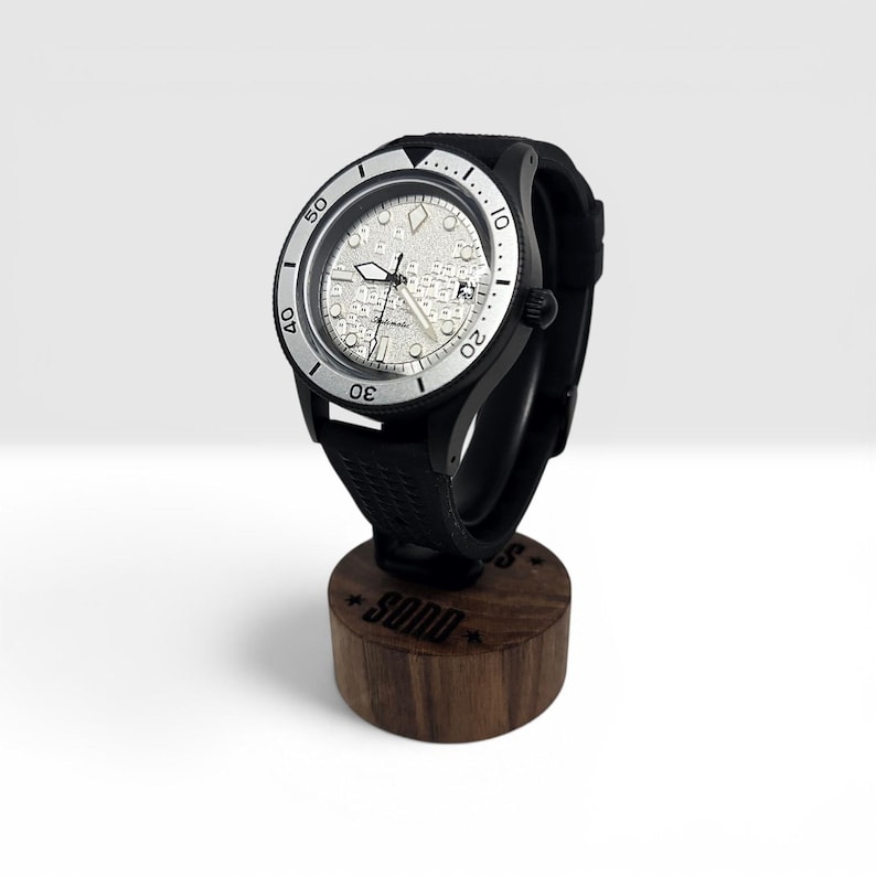 May include: An automatic wristwatch with a black case and a textured black rubber strap. The watch face features a silver-toned dial with a complex, map-like pattern, luminous hands, and a date window. The bezel is silver with black numerals.