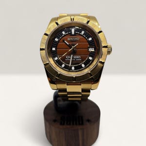 Op de afbeelding: Een goudkleurig polshorloge met een bruine wijzerplaat en zwarte accenten. Het horloge heeft de tekst "KING SEIKO AUTOMATIC 3 DAYS" en een datumweergave. Het horloge wordt gepresenteerd op een houten standaard.