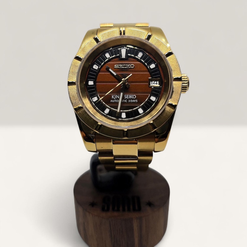 Op de afbeelding: Een goudkleurig polshorloge met een bruine wijzerplaat en zwarte accenten. Het horloge heeft de tekst "KING SEIKO AUTOMATIC 3 DAYS" en een datumweergave. Het horloge wordt gepresenteerd op een houten standaard.