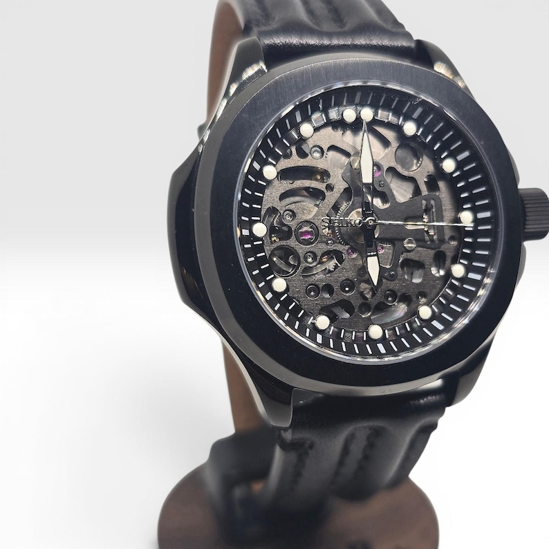 May include: A black Seiko wristwatch featuring a skeletonized dial that reveals the internal mechanical movement. The watch has a round black case, white hour markers, luminous hands, and a black leather strap with visible stitching details.