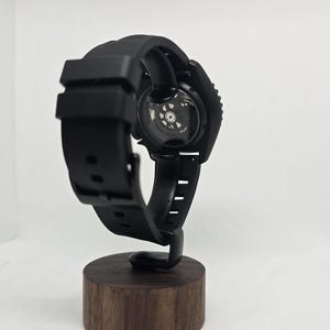 May include: A black wristwatch with a silicone strap and a circular case featuring an exhibition case back that reveals the internal mechanical movement. The watch is mounted on a cylindrical wooden stand, showcasing the back of the watch.