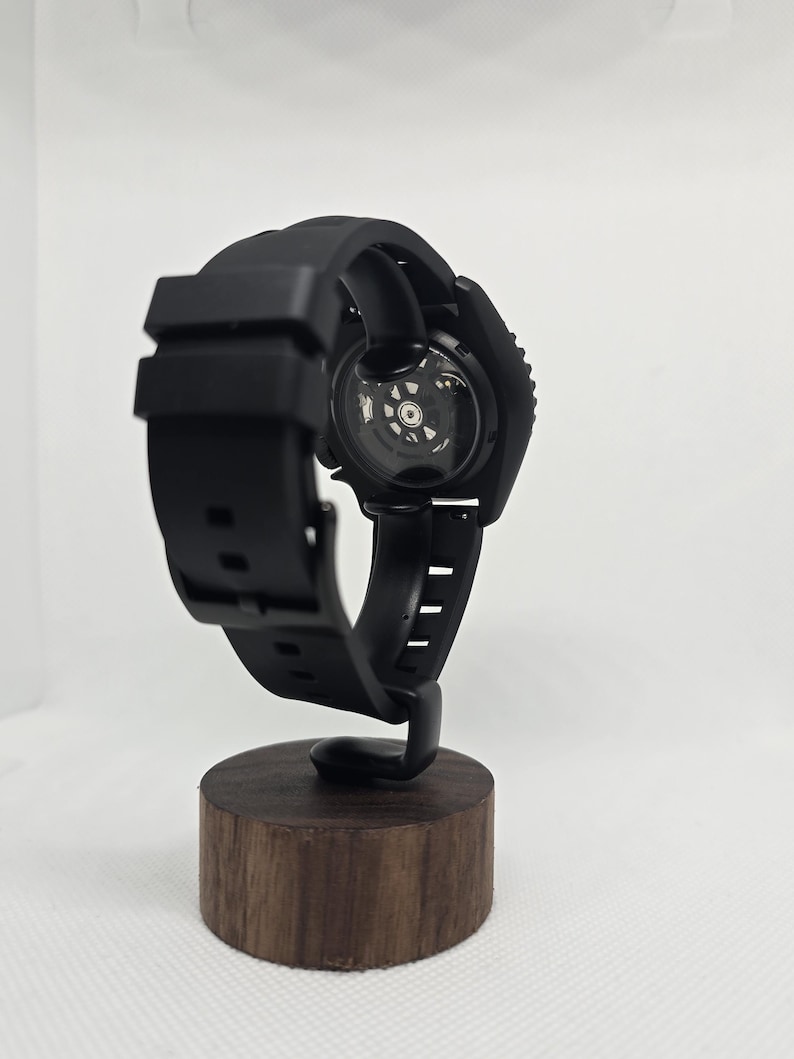 May include: A black wristwatch with a silicone strap and a circular case featuring an exhibition case back that reveals the internal mechanical movement. The watch is mounted on a cylindrical wooden stand, showcasing the back of the watch.