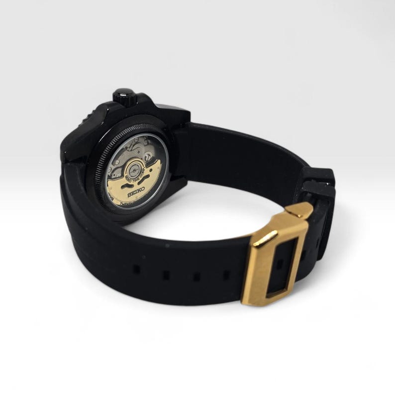 Pu&ograve; includere: Un orologio da polso nero con cinturino in silicone nero e fibbia in metallo dorato. Il fondello &egrave; trasparente e rivela il movimento meccanico interno con un rotore color oro contrassegnato dal marchio Seiko e da del testo.
