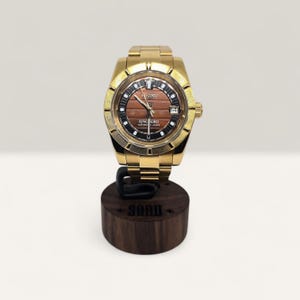 Op de afbeelding: Een goudkleurig polshorloge met een bruine en zwarte wijzerplaat, met de tekst "KING SEIKO". Het horloge wordt gepresenteerd op een donkerhouten standaard met de gravure "SONO". De band is goudkleurig. De diameter van de wijzerplaat is ongeveer 3,5 cm.