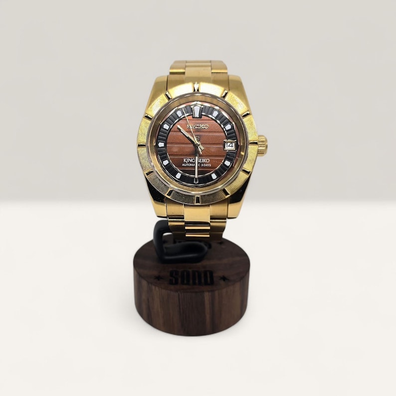 Op de afbeelding: Een goudkleurig polshorloge met een bruine en zwarte wijzerplaat, met de tekst "KING SEIKO". Het horloge wordt gepresenteerd op een donkerhouten standaard met de gravure "SONO". De band is goudkleurig. De diameter van de wijzerplaat is ongeveer 3,5 cm.