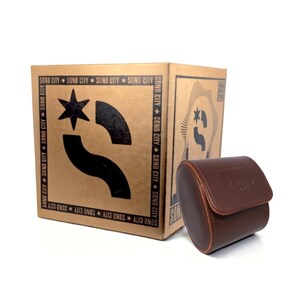 May include: A brown cylindrical leather case with a flap closure sits in front of a square cardboard box. The box is tan with black graphics, including a star, a curved shape, and the repeated text SONO CITY along the border edges.