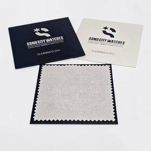 May include: A black square envelope, a white square envelope, and a light grey square cleaning cloth with pinked edges. Both envelopes feature the logo and text SOND CITY WATCHES CHICAGO-BASED WATCH CO. CLEANING CLOTH printed on them.
