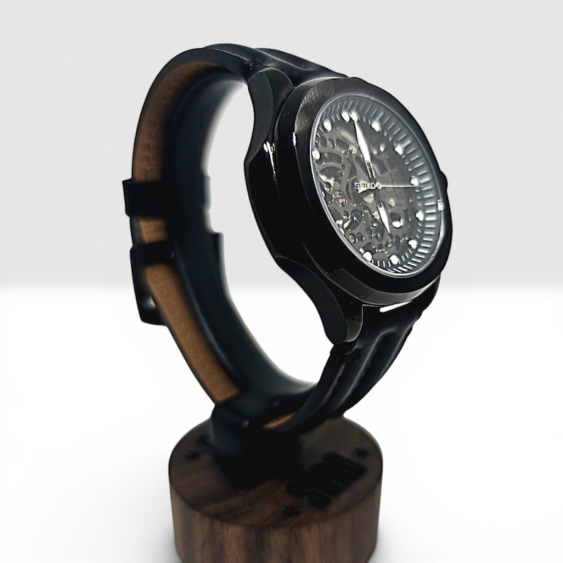 May include: A black wristwatch with a skeleton dial revealing internal mechanical components. The watch features a round black case, a black strap with a tan interior, and silver-toned hands. The word Seiko is printed on the watch face.