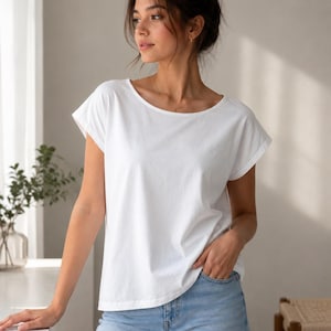 Könnte beinhalten: Ein schlichtes weißes Kurzarm-T-Shirt mit weitem Rundhalsausschnitt und umgeschlagenen Ärmeln, kombiniert mit einer hellblauen Jeans. Das T-Shirt hat eine lockere Passform mit geradem Saum auf Hüfthöhe und besteht aus einfarbigem Stoff.