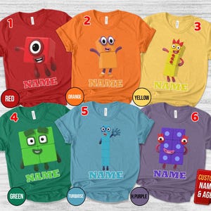 May include: Six short-sleeve t-shirts in red, orange, yellow, green, turquoise, and purple. Each shirt features a Numberblocks character graphic and the word NAME printed below. The shirts are numbered one through six in the top corners.