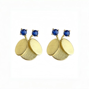 May include: A pair of gold-coloured stud earrings. Each earring features three textured, oval-shaped petals and two small, round, blue gemstones. The earrings are designed to resemble a stylized flower or insect.
