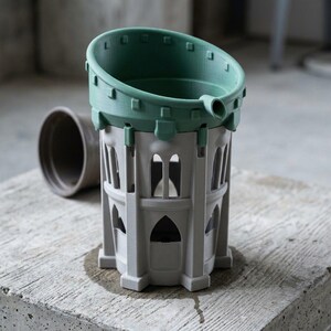 Peut inclure: Un pot de fleurs en deux parties composé d'une base gris clair avec des découpes en arc et d'une section supérieure vert sauge dotée d'un rebord circulaire et d'un petit bec verseur, au design architectural et fini mat.