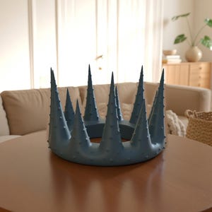Può includere: Un oggetto circolare blu opaco a forma di corona caratterizzato da molteplici punte alte e acuminate che si estendono verso l'alto da una base solida. L'intera superficie dell'oggetto è ricoperta da piccole protuberanze in rilievo, conferendogli un aspetto strutturato.