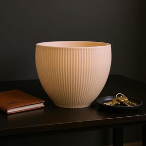 May include: A peach-colored, ribbed ceramic planter sits on a dark wooden surface. A small black dish with gold keys and a brown leather-bound notebook are also on the surface. The planter has a rounded shape and vertical grooves.