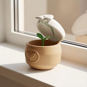 May include: A tan, bowl-shaped container with a hinged white lid that is open. Inside the container, a small green sprout with two leaves stands upright. The exterior of the tan container features a circular embossed emblem of a small plant.