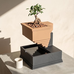 Op de afbeelding: Een beige, getrapte plantenbak met een kleine groene bonsaiboom en bruine kleikorrels, zwevend boven een donkergrijze rechthoekige bak. Een kleine, effen witte cilindrische beker staat links van de donkergrijze basis op het oppervlak.