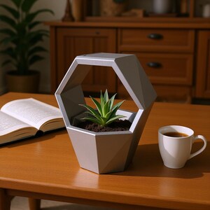 Op de afbeelding: Een zilverkleurige, geometrische, zeshoekige plantenbak met een gefacetteerd exterieurontwerp. De plantenbak bevat een kleine, groene vetplant geworteld in donkere aarde. De structuur heeft een open frame aan de bovenzijde.