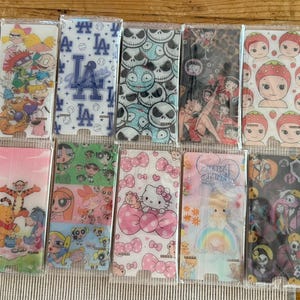 May include: A collection of fourteen rectangular plastic phone stands, each individually wrapped in clear plastic. The stands feature various printed designs including cartoon characters, sports logos, skulls, bows, and movie-themed graphics.