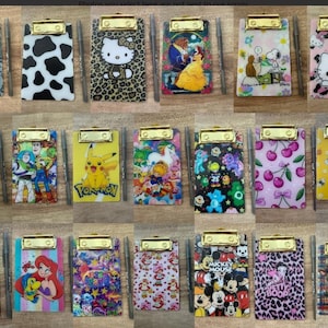 May include: A collection of eighteen rectangular clipboards with gold-tone metal clips, each featuring distinct colorful graphic prints including cartoon characters, animal patterns, and fruit designs, with a thin pen placed beside each clipboard.