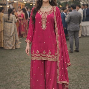 May include: A magenta-colored, embroidered salwar kameez ensemble. The outfit includes a long-sleeved tunic, wide-legged pants, and a matching scarf. Gold embroidery adorns the neckline, cuffs, and hem. The person is wearing gold jewelry and sandals.