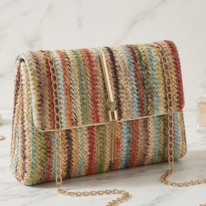 K&ouml;nnte beinhalten: Eine rechteckige Clutch mit einem gewebten Streifenmuster in Beige-, Rot-, Gr&uuml;n- und Blaut&ouml;nen. Die Tasche hat einen goldfarbenen Kettenriemen und einen goldfarbenen Stab mit Quaste. Die Clutch ist ein stilvolles Accessoire.