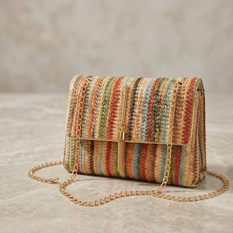 K&ouml;nnte beinhalten: Eine rechteckige Handtasche mit einem gewebten Streifenmuster in Braun-, Orange-, Gr&uuml;n- und Blaut&ouml;nen. Die Tasche hat einen goldfarbenen Kettenriemen und ein goldenes Quastendetail auf der Frontklappe. Die Tasche ist etwa 23 cm breit.