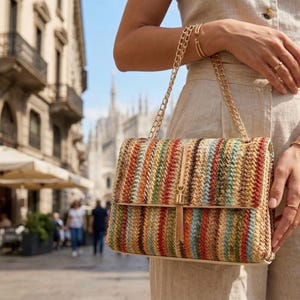 K&ouml;nnte beinhalten: Rechteckige Handtasche mit gewebtem Streifenmuster in verschiedenen Farben, darunter Rot, Orange, Gr&uuml;n und Braun. Die Tasche hat einen goldfarbenen Kettenriemen und einen Quastenanh&auml;nger. Im Hintergrund ist eine Stadtstra&szlig;e zu sehen.