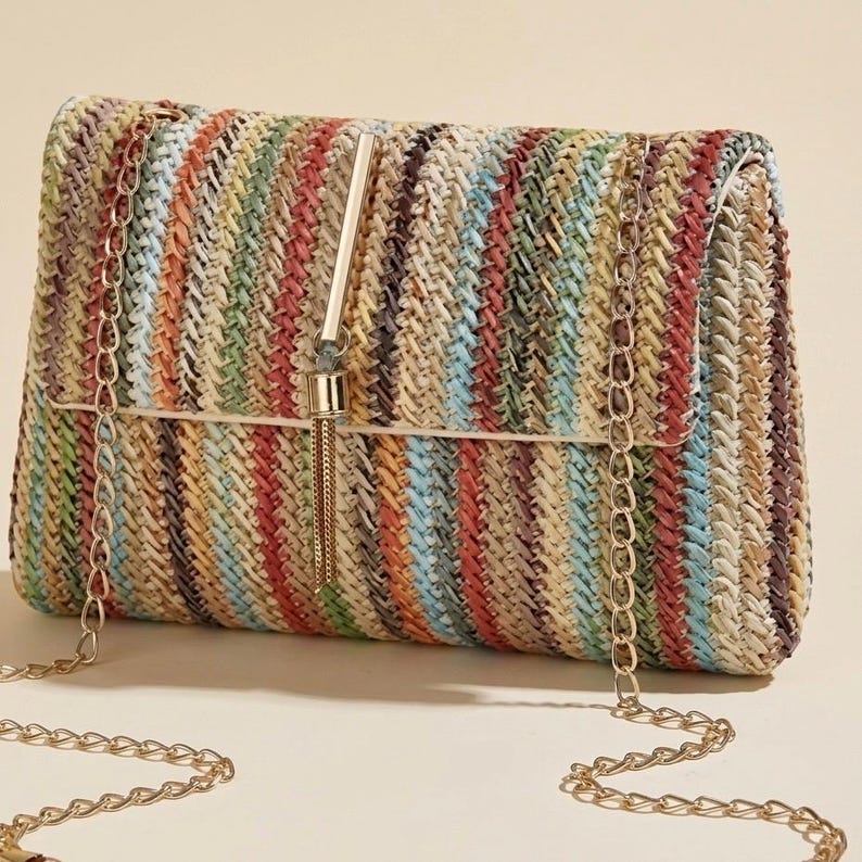 K&ouml;nnte beinhalten: Eine rechteckige Clutch mit einem gewebten, gestreiften Muster in verschiedenen Farben, darunter Rot, Blau, Gr&uuml;n und Gelb. Die Clutch hat einen goldenen Kettenriemen und ein Quastendetail. Die Tasche besteht aus einem stroh&auml;hnlichen Material.