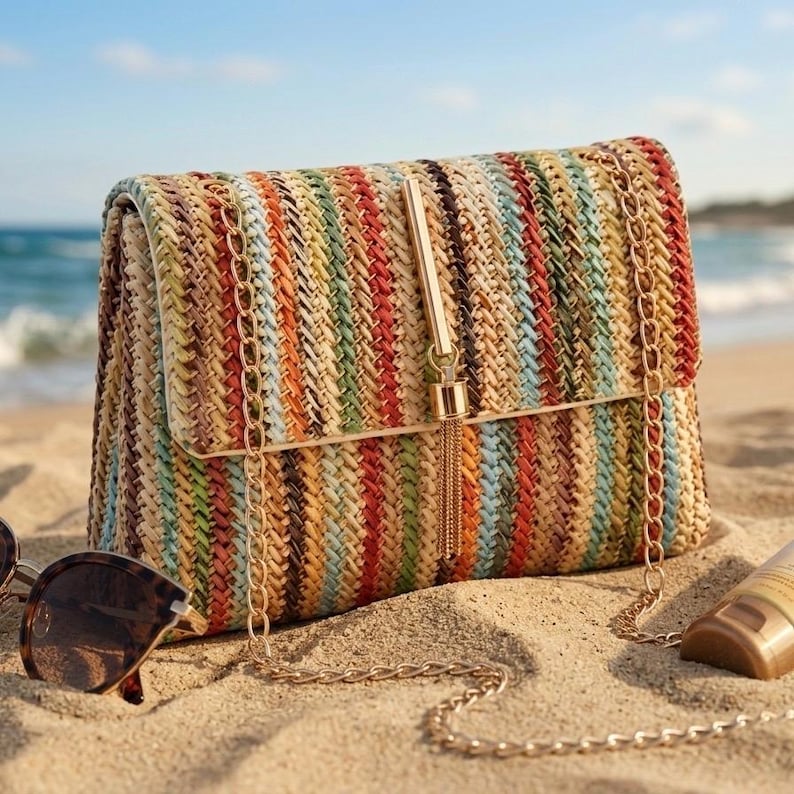 K&ouml;nnte beinhalten: Mehrfarbige gewebte Clutch mit goldener Kette und Quasten. Die Tasche hat einen Klappverschluss und liegt an einem Sandstrand. Sonnenbrille und Sonnencreme sind ebenfalls zu sehen.