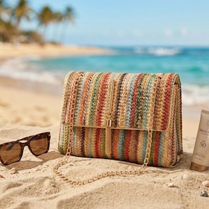 K&ouml;nnte beinhalten: Eine mehrfarbige gewebte Handtasche mit goldener Kette und Quaste, die an einem Sandstrand neben Sonnenbrille und Sonnencreme liegt. Die Tasche hat vertikale Streifen in Orange-, Gr&uuml;n- und Blaut&ouml;nen.