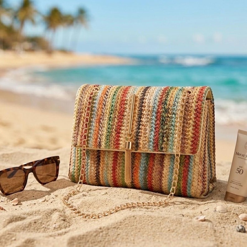 K&ouml;nnte beinhalten: Eine mehrfarbige gewebte Handtasche mit goldener Kette und Quaste, die an einem Sandstrand neben Sonnenbrille und Sonnencreme liegt. Die Tasche hat vertikale Streifen in Orange-, Gr&uuml;n- und Blaut&ouml;nen.