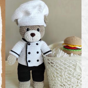 May include: A crocheted teddy bear dressed as a chef, wearing a white chef's hat, jacket, and black pants. A crocheted hamburger sits in a white basket. The bear has a brown nose and is standing on a light-colored surface.